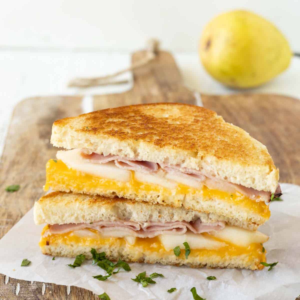 A pear grilled cheese sandwich with pear slices and deli ham cut in half and stacked on a wooden cutting board.