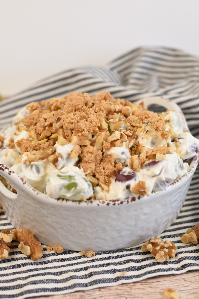 Whtie serving bowl on a black and white striped napkin with white grape salad and a streusel topping.