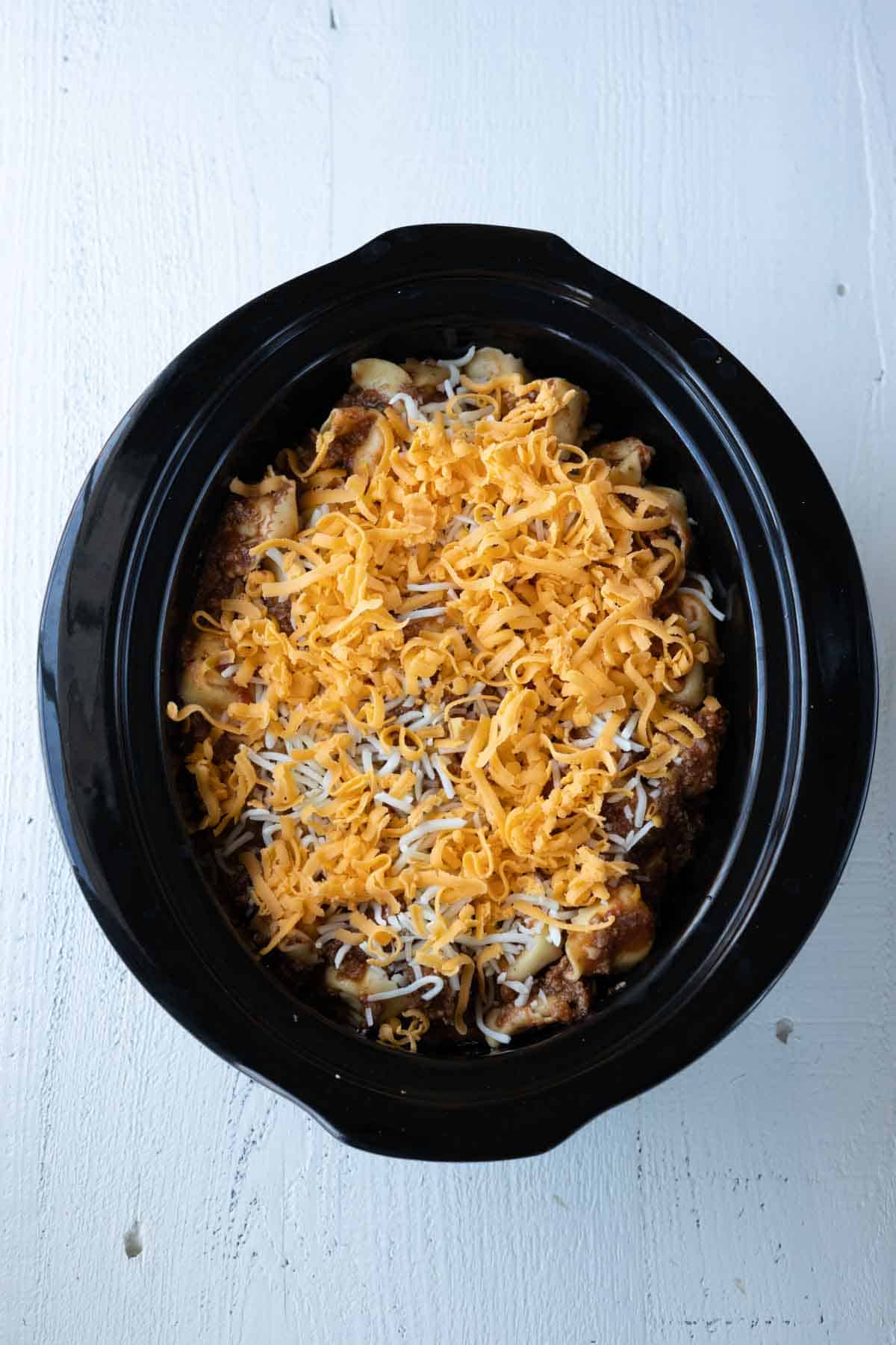 A slow cooker with beef and tortellini topped with grated cheddar cheese in a slow cooker.