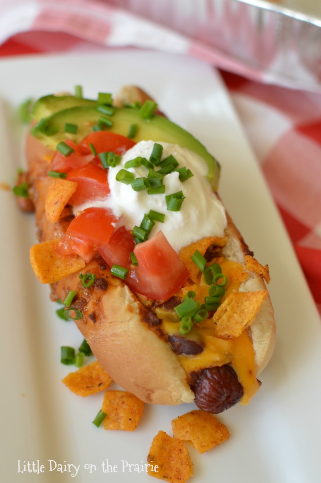 a hotdog in a bun topped with chili, diced tomatoes, sour cream, and avocados