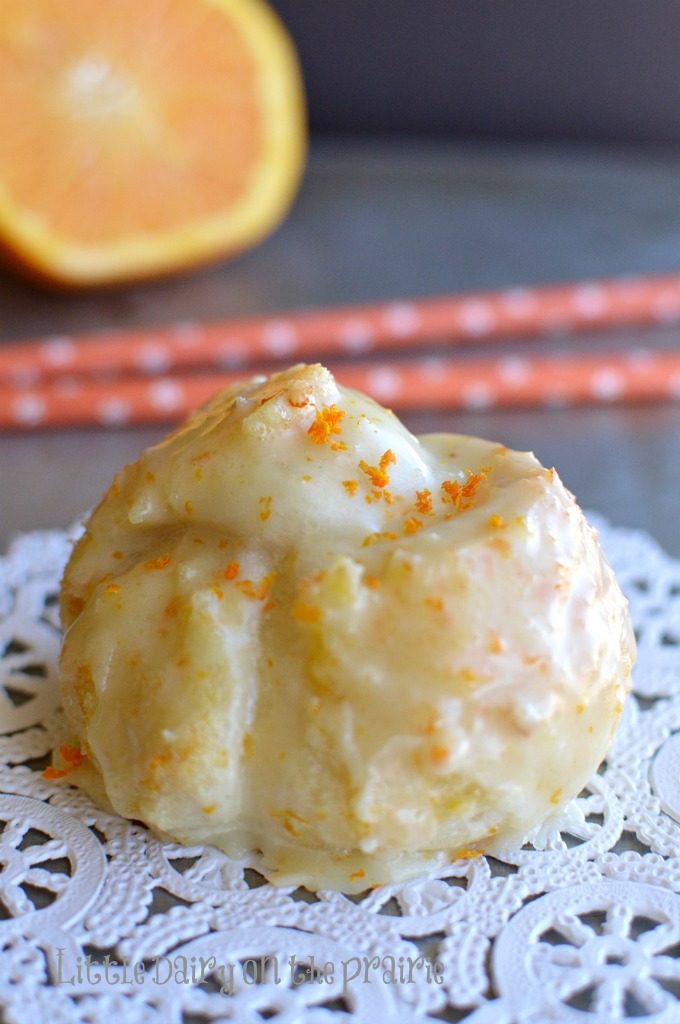 A baked orange knot glazed with orange icing, on a doilly, and two orange straws in the background