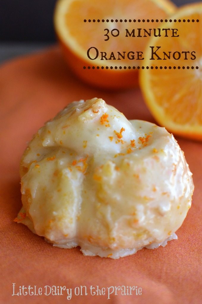 Orange knots glazed with icing, on an orange napkin with sliced oranges in the back