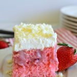 A square of pink cake with holes poked in it, then filled with strawberry and cream filling, and topped with whipped cream layer.