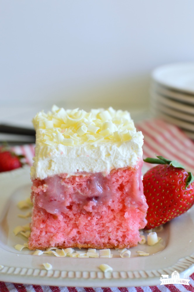 A square of pink cake with holes poked in it, then filled with strawberry and cream filling, and topped with whipped cream layer.