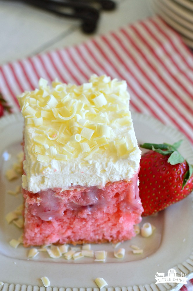 pink strawberry poke cake with holes filled with sweetened condensed milk and topped with a layer of white whipped cream and sprinkled with white chocolate curls. Little Dairy on the Prairie
