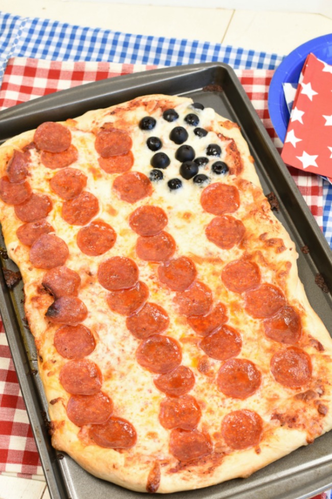 Pepperoni pizza that looks like and American flag with olives in the corner and sitting on a red and white and blue and white gingham table cloth.