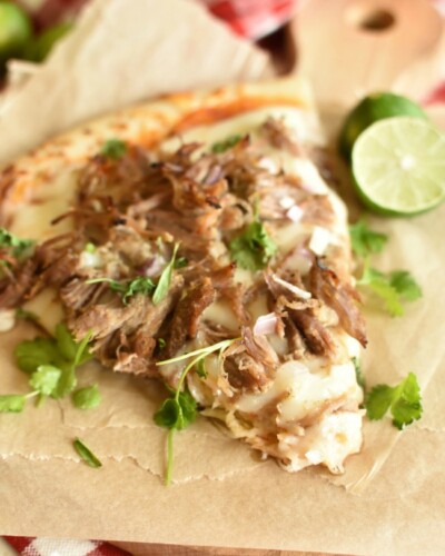 a slice of pizza topped with shredded pork and cilantro on top of a brown paper and a lime half