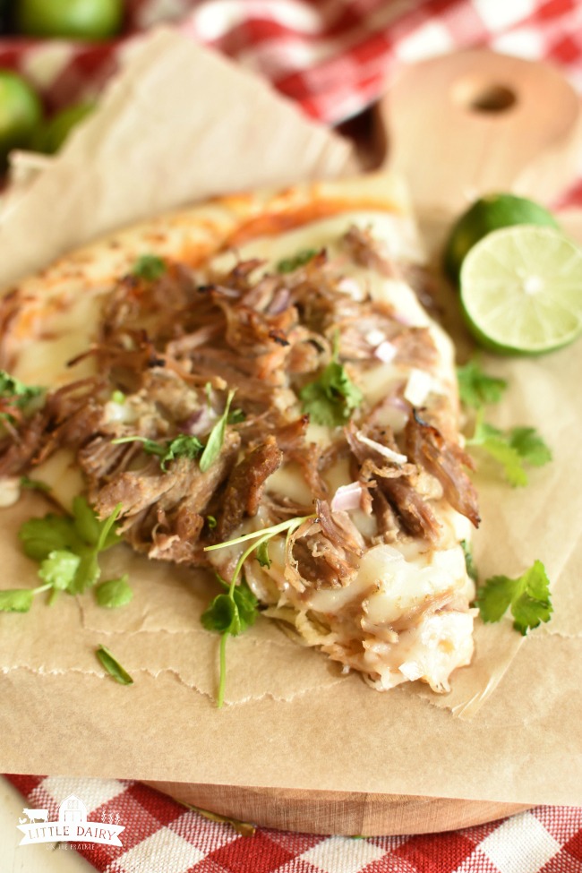 a slice of pizza topped with shredded pork and cilantro on top of a brown paper and a lime half