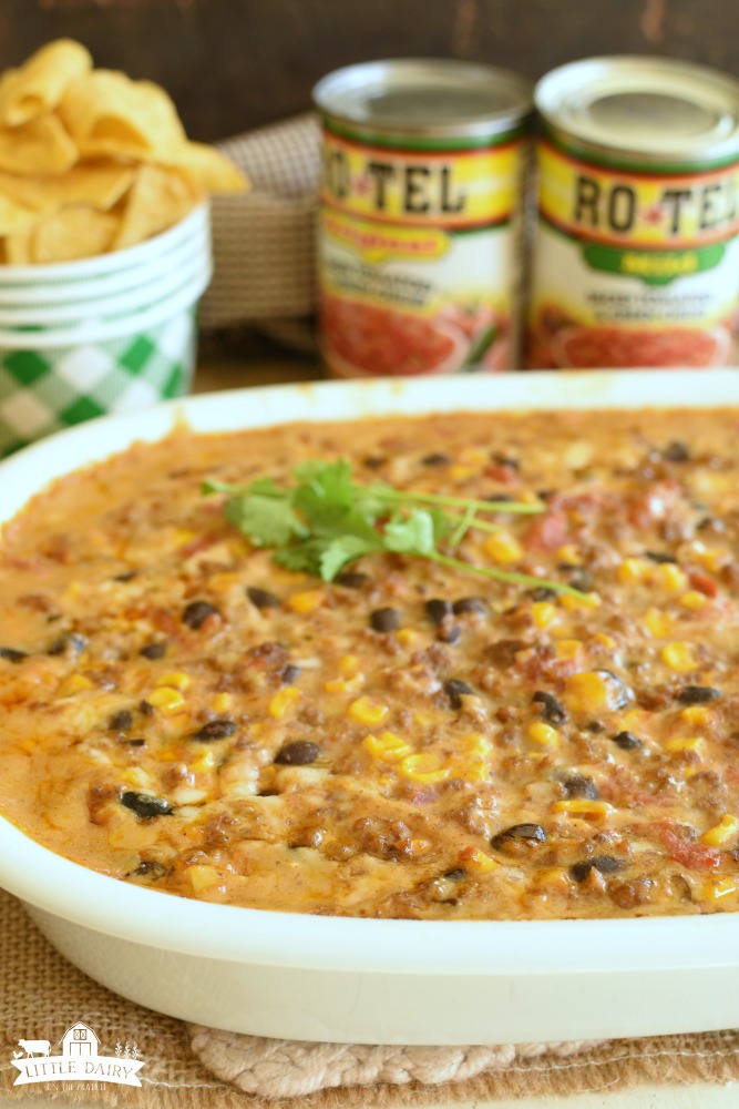 A cheesy Mexican corn dip with black beans, corn, and tomatoes in a white casserole dish.