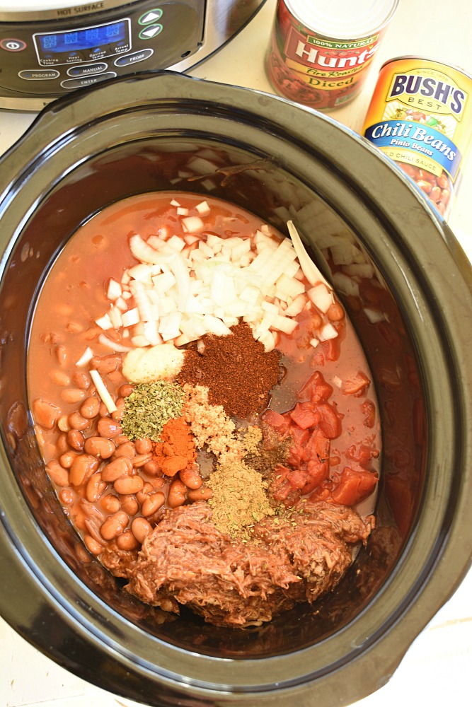 a black slow cooker filled with beans, pulled pork, tomatoes, spices, and onions not stirred together