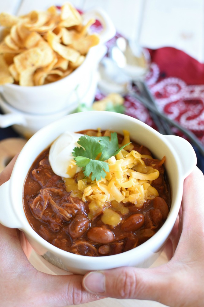 a white bowl filled with a red chili and topped with shredded cheddar cheese and sour cream