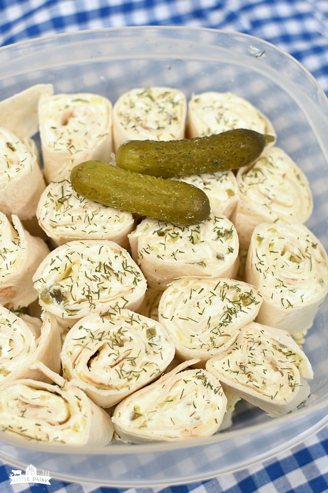 flour tortillas pinwheels with cream cheese and dill weed, two dill pickles sitting on top. In a clear plastic container, sitting on a blue and white checkered cloth