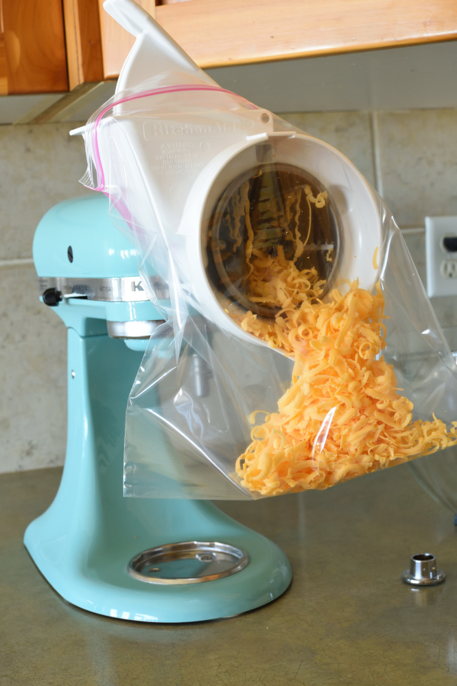 A light blue ktichenaid mixer with a food processor attaachment grating cheese into a gallon sealable bag