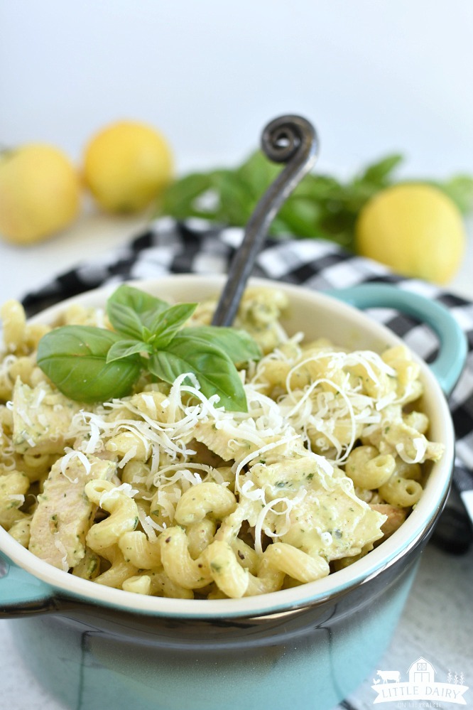 pesto pasta in a bowl with basil on top of it and a black spoon in it with lemons in the background on a black and white checkered napkin