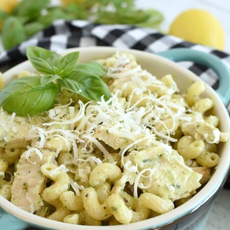 Pasta with grated parmesan cheese and grilled chicken on top of it in a blue dish, a black and white checkered napkin, lemons and basil in the background