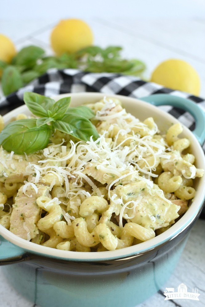 Pasta with grated parmesan cheese and grilled chicken on top of it in a blue dish, a black and white checkered napkin, lemons and basil in the background