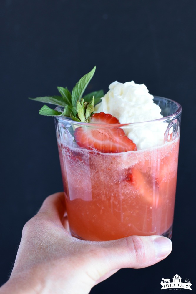 A hand jp;domg a red drink with strawberries floating on top, whipped cream, and a mint sprig