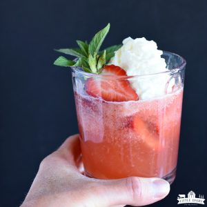 A red drink in a clear glass with strawberries floating on top and whipping cream