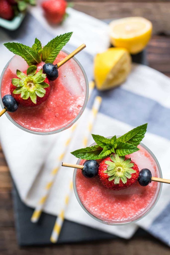 two glasses of strawberry coolers with a toothpick skewered with strawberries, blueberries, and mint