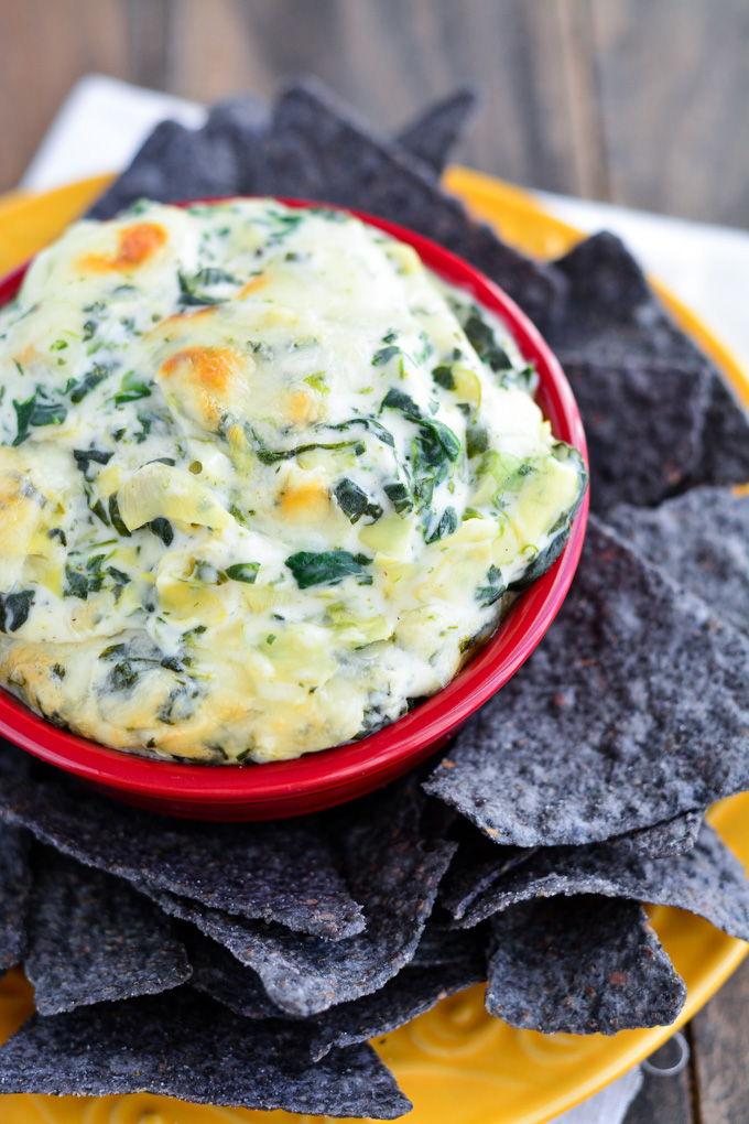 a red bowl filled with cream spinach and artichoke dip topped with cheese and served with blue corn chips