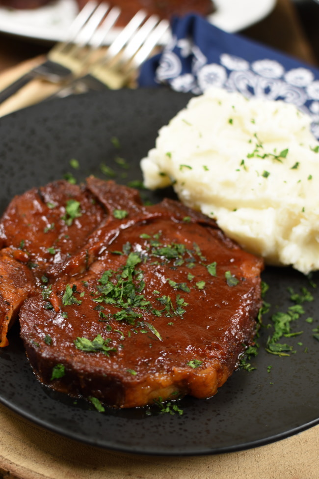 A cooked pork chop basted in BBQ sauce on a black plate with mashed potatoes
