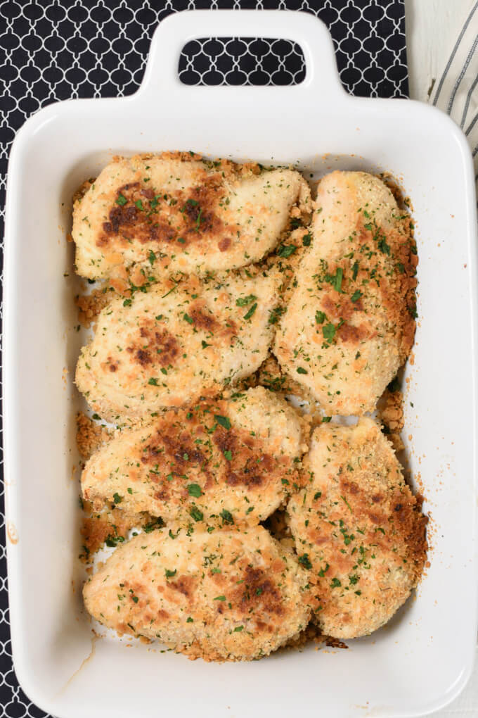 A white baking dish with golden brown baked chicken breasts breaded and sprinkled with chopped green parsley