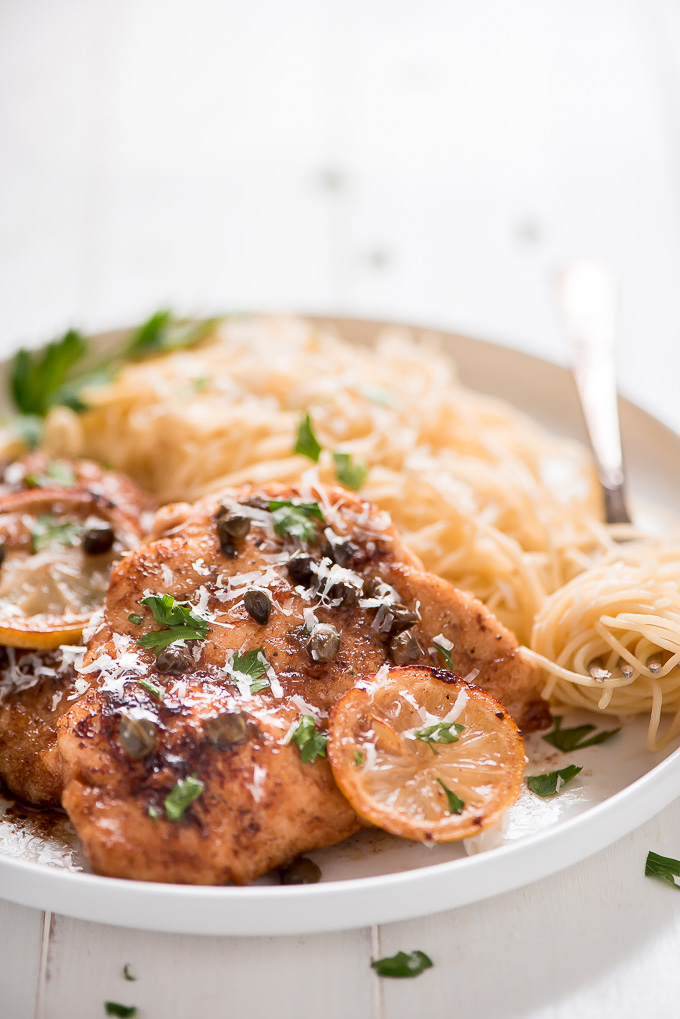 A breaded and baked chicken breast topped with sliced lemons, shredded parmesan cheese, capers, and parsley on a plate with angel hair pasta