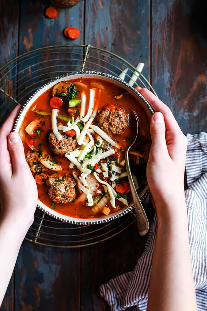 a hand holding a bowl of soup with meatballs, grated mozzarella cheese, vegetables, and spoon