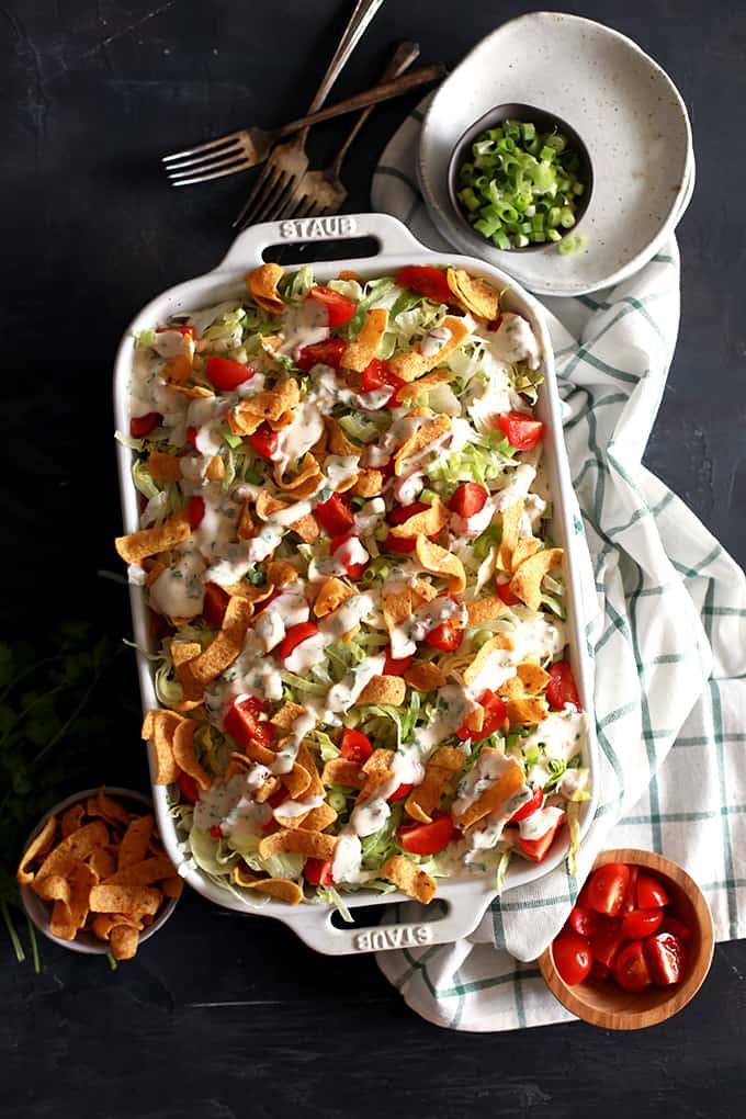 a white baking dish with taco pizza, topped with shredded lettuce, corn chips, diced tomatoes, and drizzled with sour cream