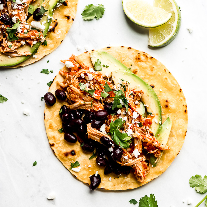 a corn tortilla shell topped with shredded chicken, avocado slices, beans, and white cheese. lime wedges