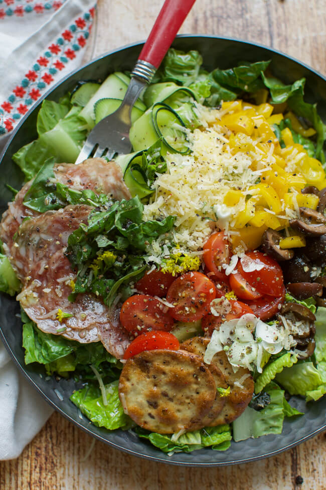 a shallow black bowl filled with salad made with peppers, tomatoes, olives, cheese, crackers, and salami