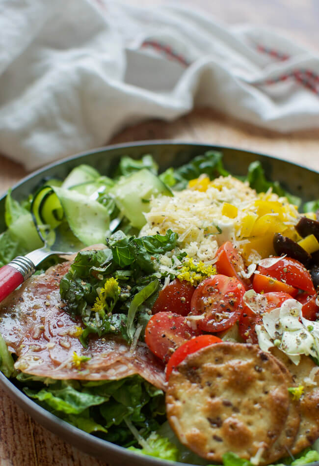 a shallow black bowl filled with salad made with peppers, tomatoes, olives, cheese, crackers, and salami