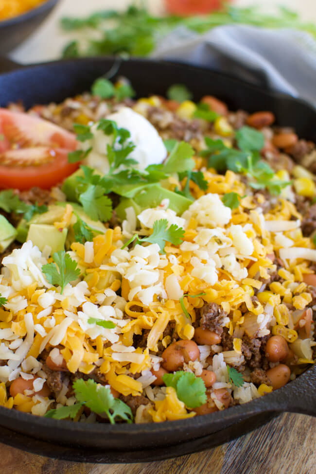 a cast iron skillet with fried hamburger, beans, corn, grated cheese, cilantro and avocados