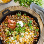 a cast iron skillet with fried hamburger, beans, corn, grated cheese, cilantro and avocados