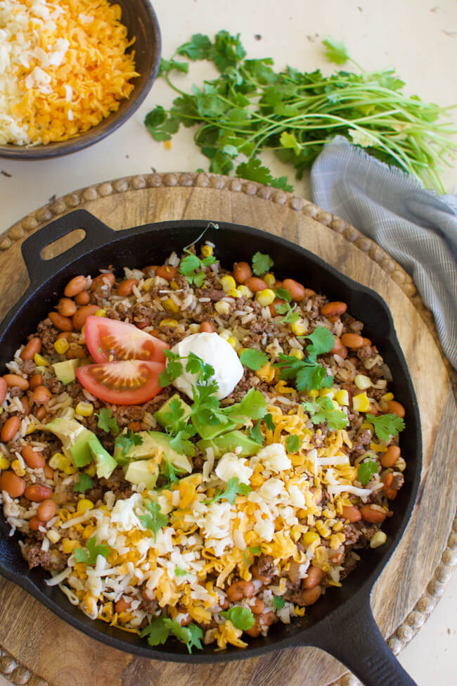 a cast iron skillet with fried hamburger, beans, corn, grated cheese, cilantro and avocados