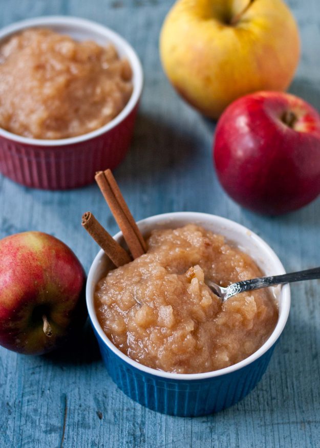 a blue ramekin filled with chunky applesauce, garnished with cinnamon sticks and a spoon. Red apples in the background.