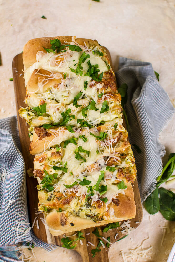 an flat lay image of pull apart spinach and artichoke bread on a wooden cutting board with parsley and grated cheese