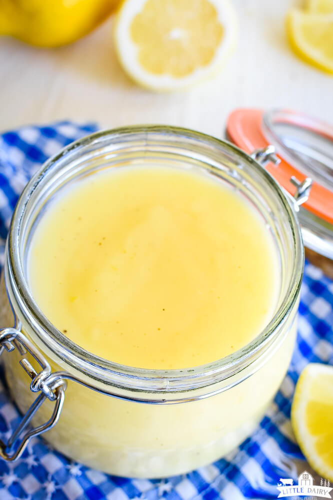 a close up shot of pale yellow lemon curd in a glass jar over a blue and white gingham napkin. Little Dairy on the Prairie