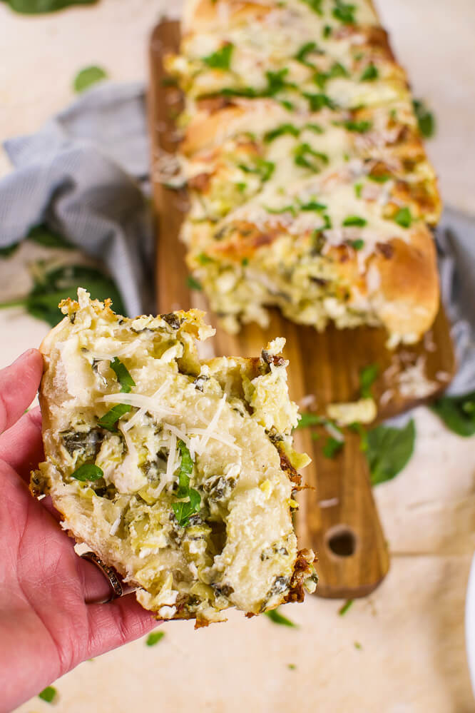 A hand holding a slice of cheesy spinach artichoke bread with cheese and chopped parsley with a bread loaf in the background