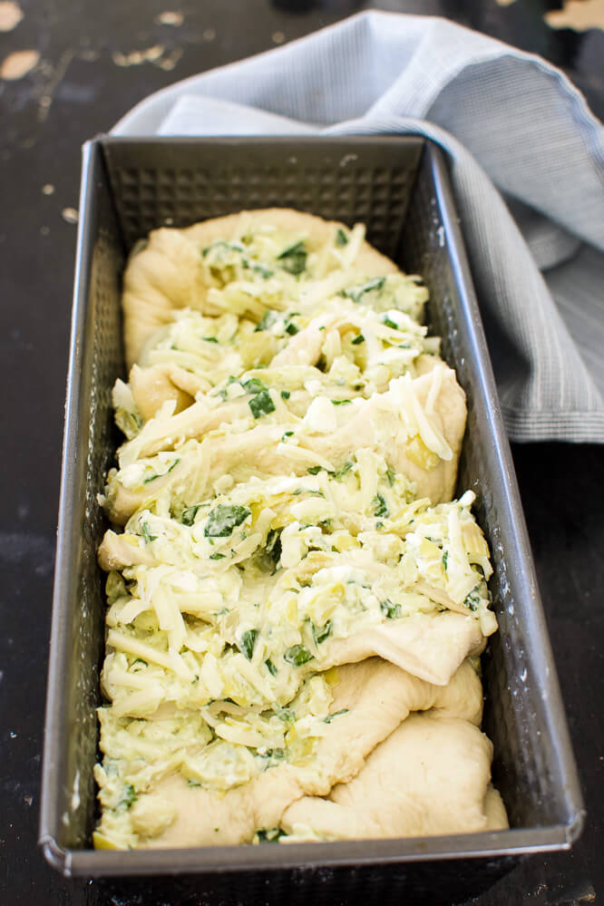 a breSquares of bread dough and layers of spinach artichoke dip stacked in a black bread pan