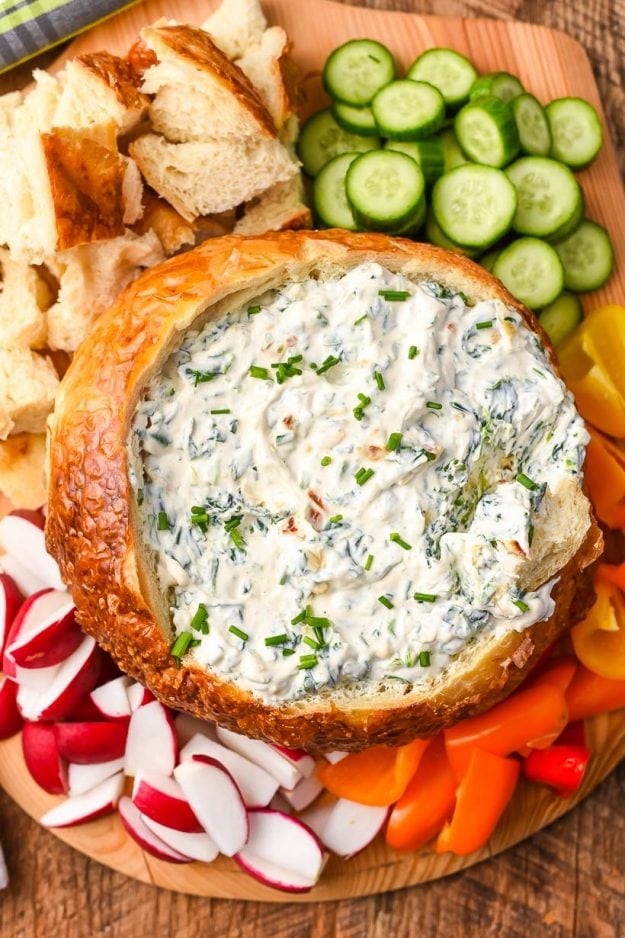 a bread bowl filled with creamy spinach dip surrounded by cucumbers, carrots, peppers, and radishes