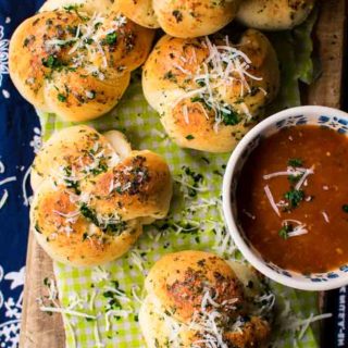 a wooden board with green and white checked piece of parchment paper with garlic knots topped with parmesan cheeseand marinara sauce