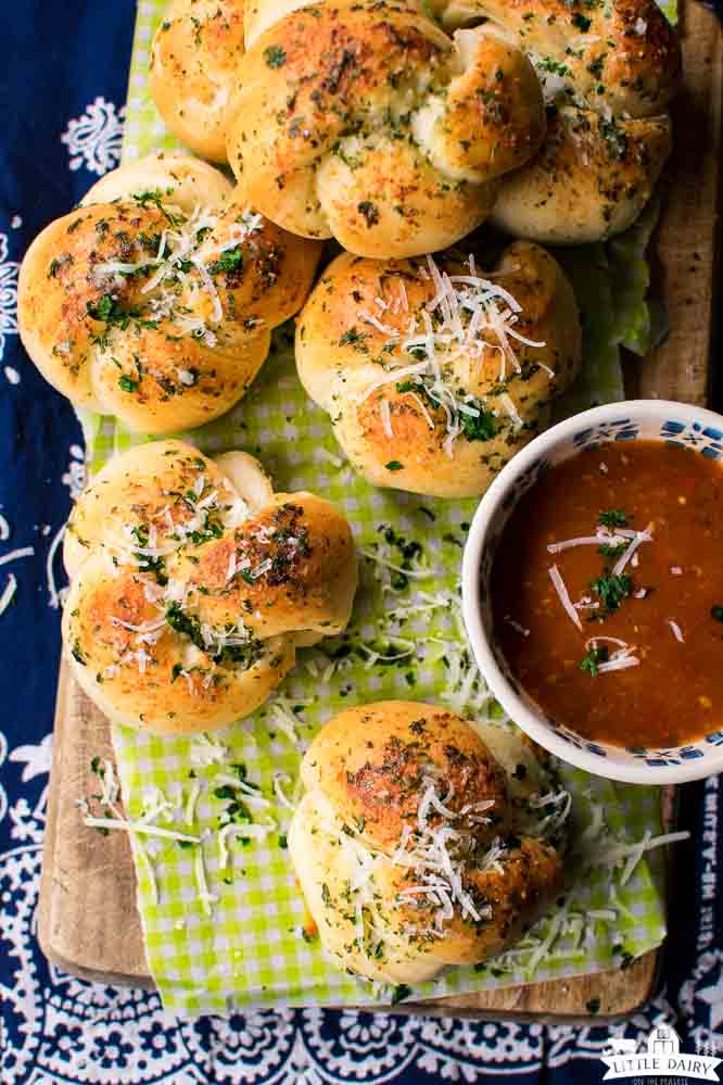 a wooden board with green and white checked piece of parchment paper with garlic knots topped with parmesan cheeseand marinara sauce
