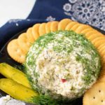 a cheese ball made with dill pickle relish and covered with fresh dill weed, surrounded by butter crackers and a pickle slice.