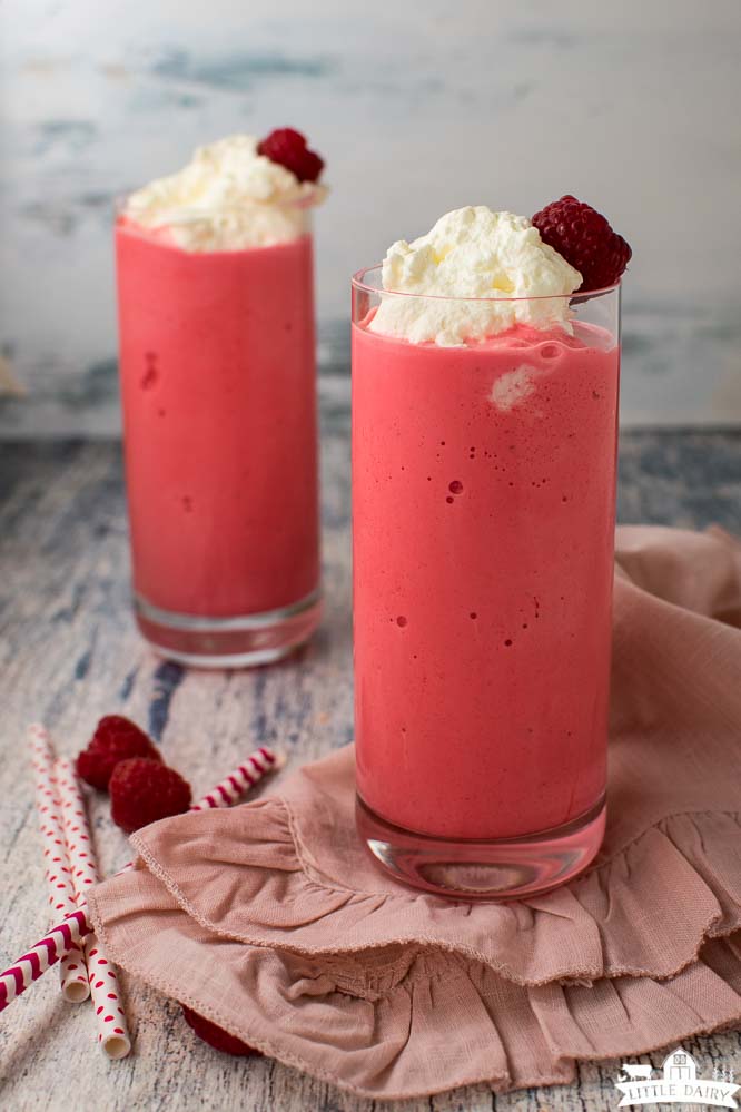 two glasses filled with bright pink milkshakes, topped with whipped cream and a raspberry