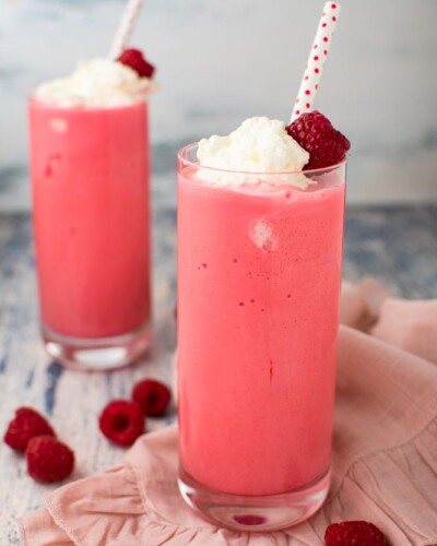 two glasses filled with fruity milkshakes, topped with whipped cream and a fresh raspberry