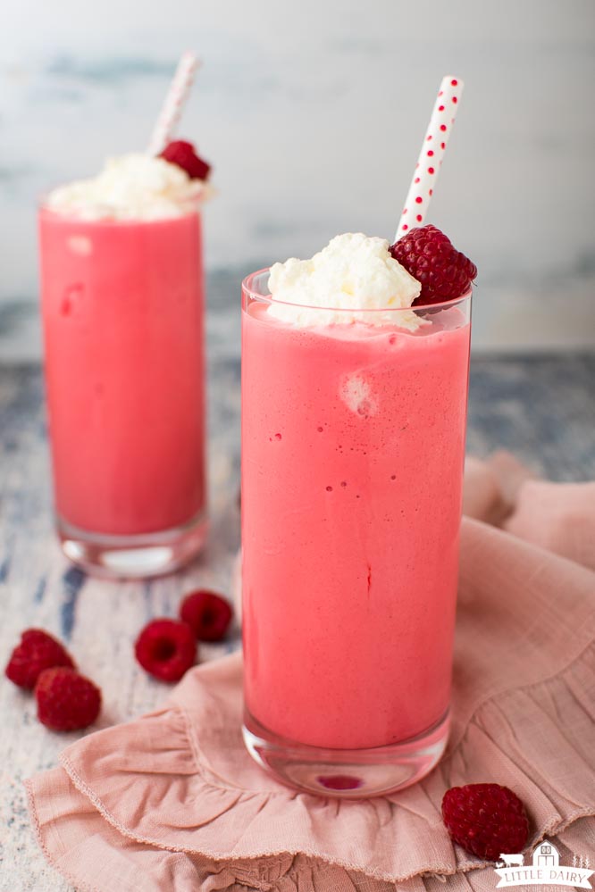 two glasses filled with fruity milkshakes, topped with whipped cream and a fresh raspberry