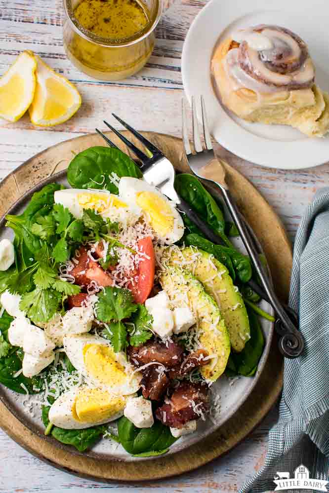 a green salad on a plate with a wooden charger, forks, lemon wedges, salad dressing and a cinnamon roll