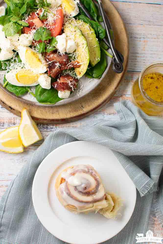 a baked and frosted cinnamon roll and a plate with a savory breakfast salad loaded with vegetables