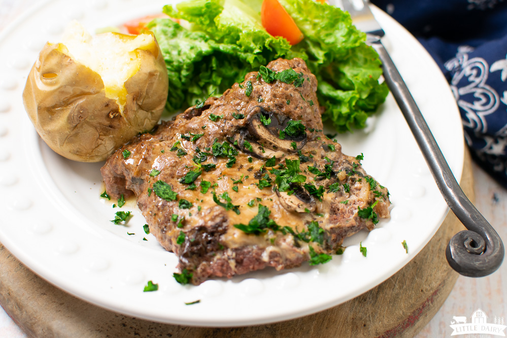 A white plate with cooked cube steak in gravy and green salad and a potato